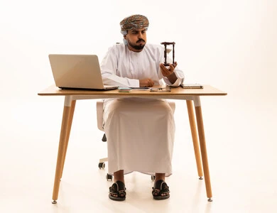 Omani Man with Hourglass and Laptop at Office Desk