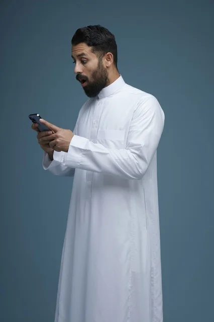 رجل سعودي بالثوب التقليدي ينظر بهاتفه الذكي بدهشة