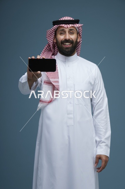 رجل سعودي يبتسم ويمسك هاتف ذكي بالثوب والشماغ