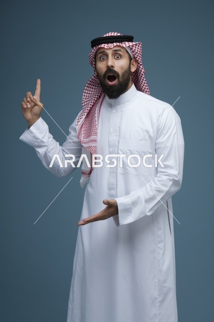رجل سعودي متفاجئ بالزي التقليدي يشير بيده أمام خلفية سادة