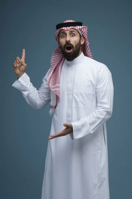 رجل سعودي متفاجئ بالزي التقليدي يشير بيده أمام خلفية سادة