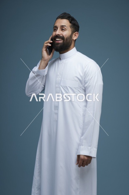 رجل سعودي مبتسم يتحدث عبر الهاتف الذكي بالثوب الأبيض
