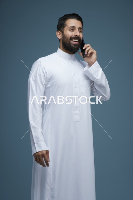 رجل سعودي مبتسم بالثوب الأبيض يتحدث بالهاتف الذكي
