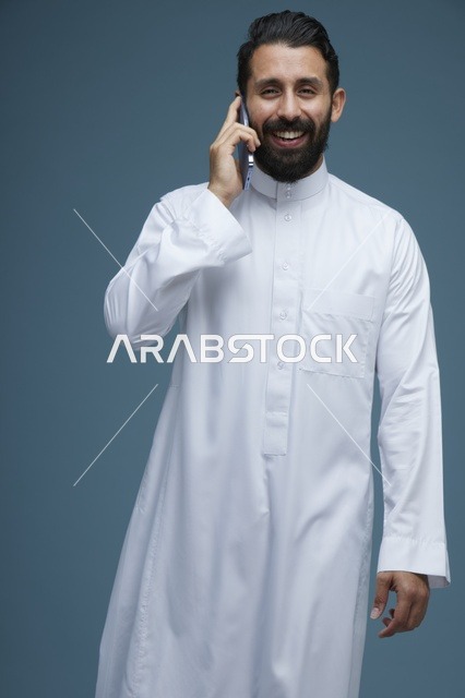 رجل سعودي مبتسم يتحدث عبر الهاتف الذكي بالثوب الأبيض