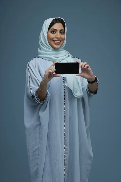 امرأة سعودية تحمل هاتفاً ذكياً بوضعية أفقية في استوديو