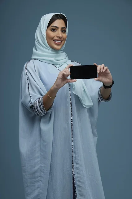 امرأة سعودية تحمل هاتفاً ذكياً بخلفية استوديو زرقاء