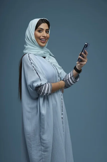 امرأة سعودية تحمل هاتف ذكي بعباءة تقليدية خلفية زرقاء
