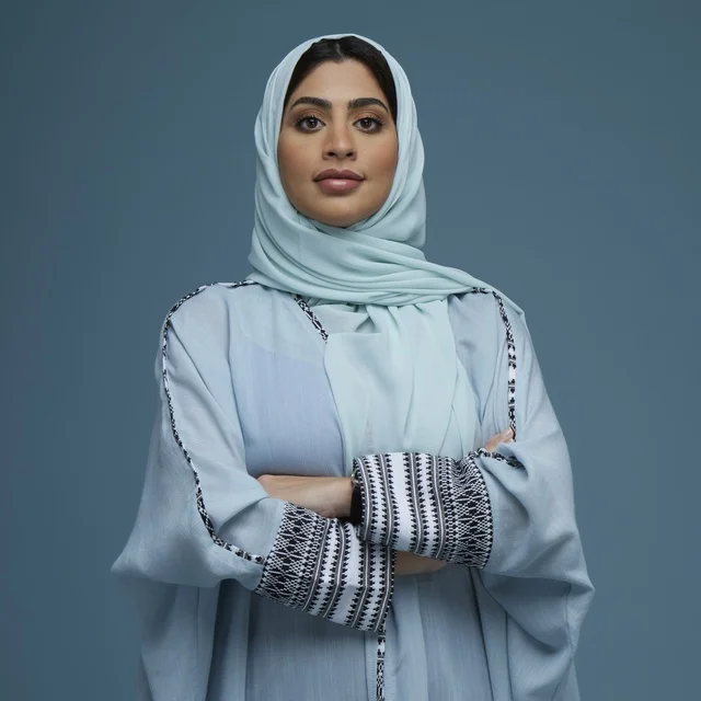 امرأة سعودية بعباءة وحجاب أزرق تقف مكتوفة اليدين