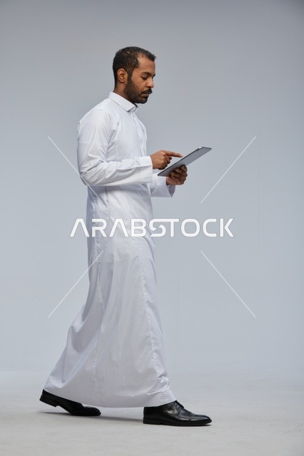 رجل سعودي بالثوب الأبيض يمشي ويستخدم تابلت
