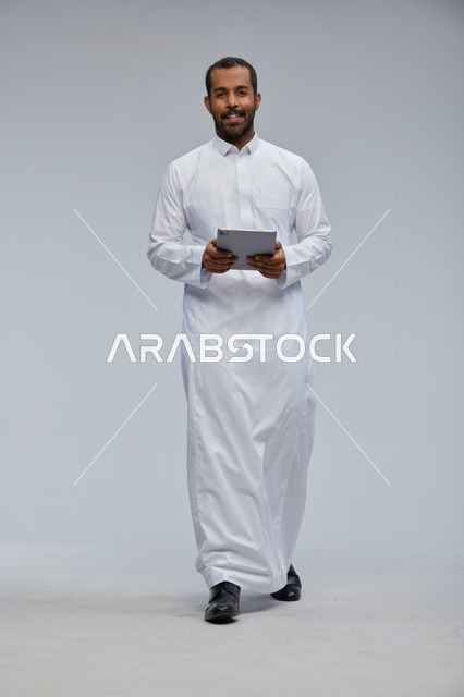 رجل سعودي بالثوب الأبيض يمسك بجهاز لوحي خلفية رمادية