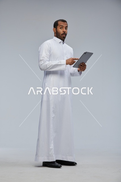 رجل سعودي بالثوب الأبيض يستخدم جهاز لوحي بتعبير مندهش