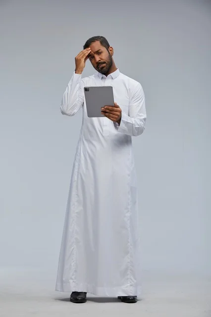 رجل سعودي بالثوب الأبيض يستخدم جهاز لوحي بتعبير قلق