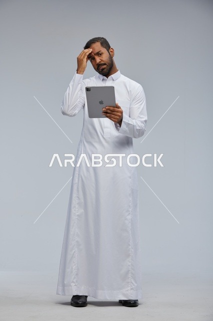 رجل سعودي بالثوب الأبيض يستخدم تابلت بتعبير حائر