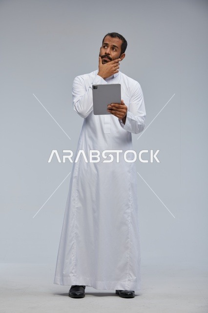 رجل سعودي بالثوب الأبيض يفكر ويستخدم جهاز لوحي