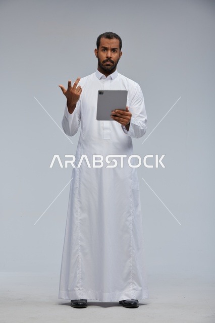 رجل سعودي بالثوب الأبيض يستخدم جهازا لوحيا بخلفية رمادية