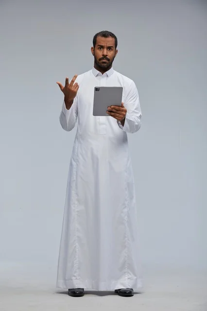 رجل سعودي بالثوب الأبيض يستخدم جهازا لوحيا بخلفية رمادية