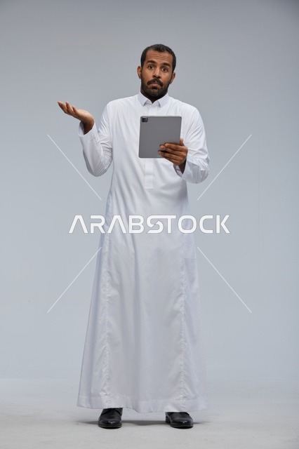 رجل سعودي بالثوب يحمل جهاز لوحي مندهش في الاستوديو