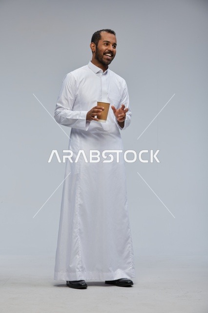 رجل سعودي مبتسم بالثوب التقليدي يحمل كوب قهوة بالاستوديو