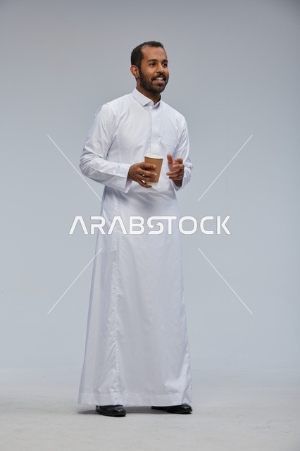 رجل سعودي مبتسم بالثوب الأبيض يحمل كوب قهوة