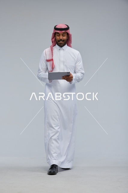 رجل سعودي يستخدم جهازاً لوحياً بالزي التقليدي يمشي