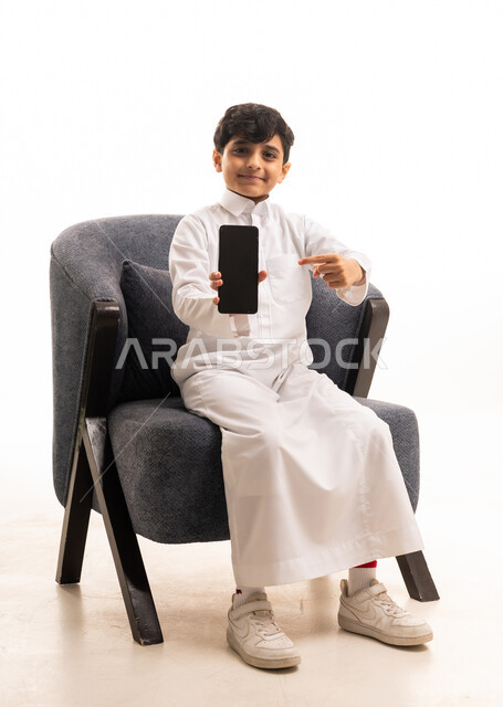 طفل سعودي بالثوب يحمل هاتف ذكي على كرسي