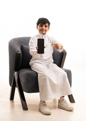 طفل سعودي بالثوب يحمل هاتف ذكي على كرسي