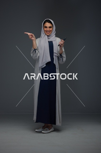 امرأة سعودية تشير للأعلى وتحمل هاتفاً ذكياً في استوديو