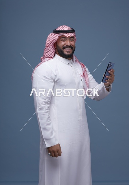 رجل سعودي مبتسم يستخدم الهاتف الذكي بالزي التقليدي