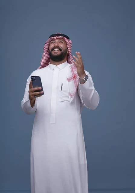 رجل سعودي مبتسم يرتدي الزي التقليدي مع هاتف ذكي