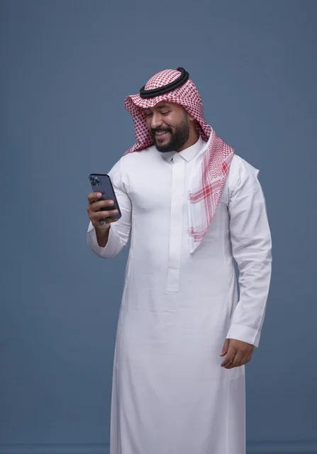 رجل سعودي بالزي التقليدي يبتسم ويستخدم الهاتف الذكي