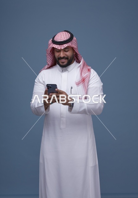 رجل سعودي مبتسم يستخدم الهاتف الذكي بالزي التقليدي