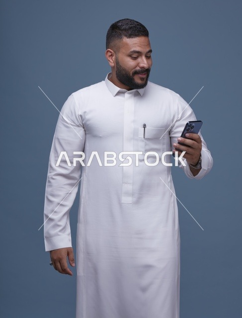 رجل سعودي بالثوب الأبيض يستخدم الهاتف الذكي بخلفية زرقاء