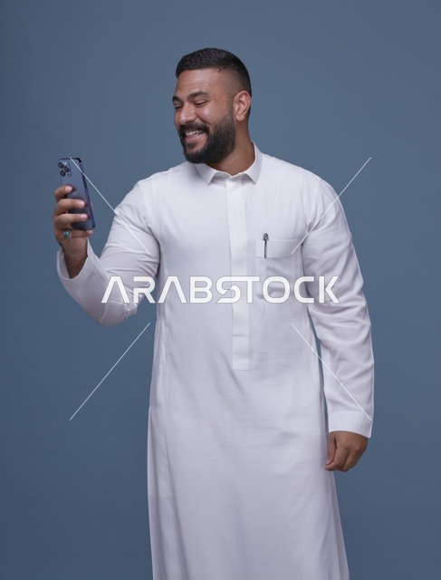 رجل سعودي يرتدي الثوب الأبيض يستخدم الهاتف الذكي بخلفية زرقاء