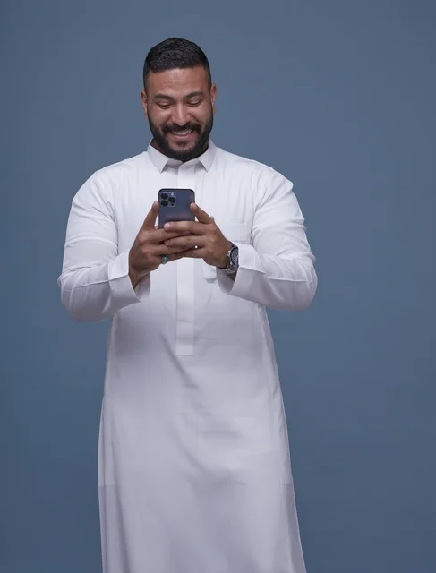 Saudi Man in White Thobe Using Smartphone on Blue Backdrop
