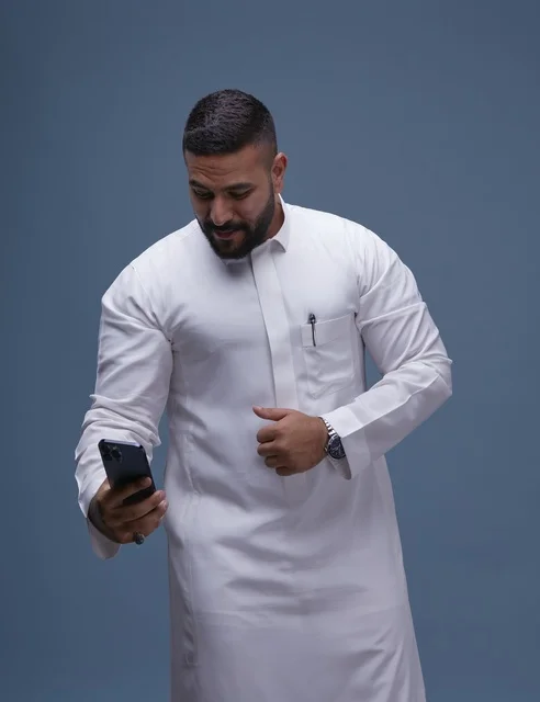 رجل سعودي مبتسم بالثوب الأبيض يلتقط سيلفي بالهاتف