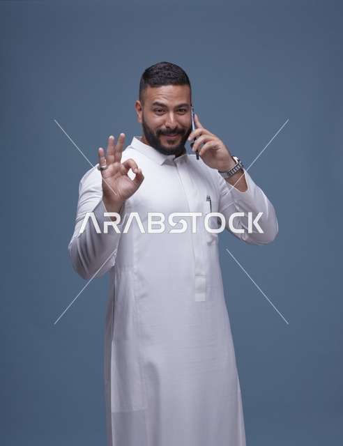 رجل سعودي بالثوب الأبيض يتحدث بالهاتف الذكي مع إشارة التمام