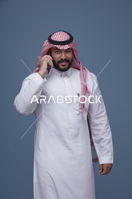 رجل سعودي مبتسم يتحدث عبر الهاتف الذكي بالزي التقليدي