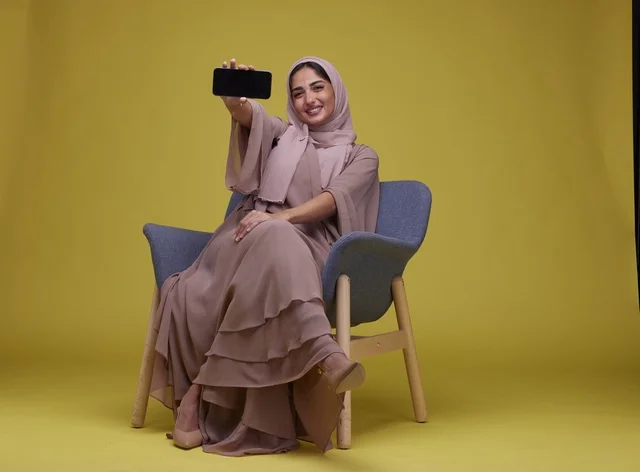 Saudi Woman Holding Smartphone in Armchair Yellow Background