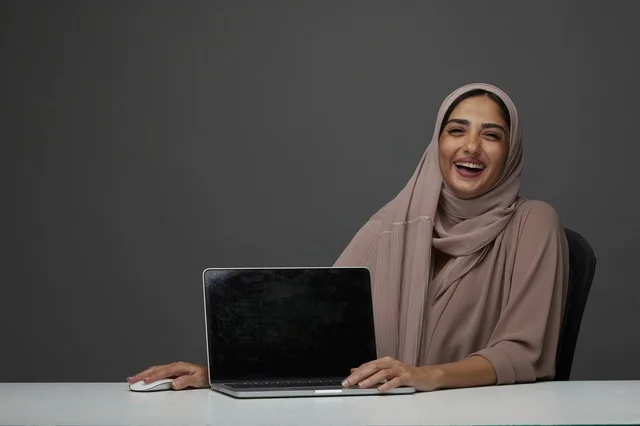 سيدة أعمال سعودية تضحك مع لابتوب في المكتب بخلفية رمادية سيدة أعمال سعودية تضحك مع لابتوب في المكتب بخلفية رمادية