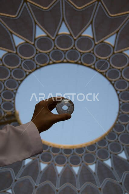 صورة جمالية مقربة ليد رجل عربي خليجي يحمل بيده مجسم على  شكل عدسة أسفل قبة ساحة الوصل في اكسبو دبي ، معلم حضاري في قلب مدينة الابداع ، تحفة معمارية عالمية ، قبة فولاذية مضيئة تجمع تحتها آلاف الاشخاص و الافكار من جميع العالم ، معرض إكسبو دبي 2022 
