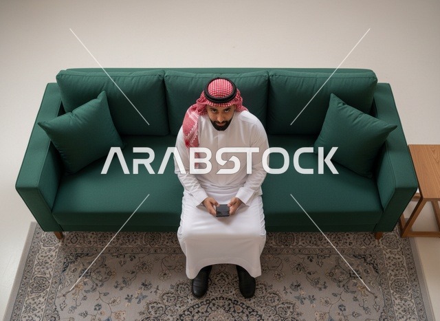 رجل سعودي بالثوب التقليدي يستخدم الجوال على الأريكة