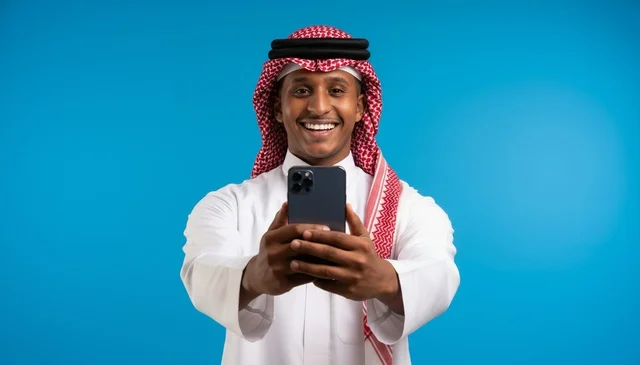 رجل سعودي مبتسم يحمل هاتف ذكي أمام خلفية زرقاء