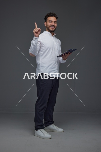 رجل سعودي يحمل جهازاً لوحياً ويشير للأعلى بخلفية رمادية