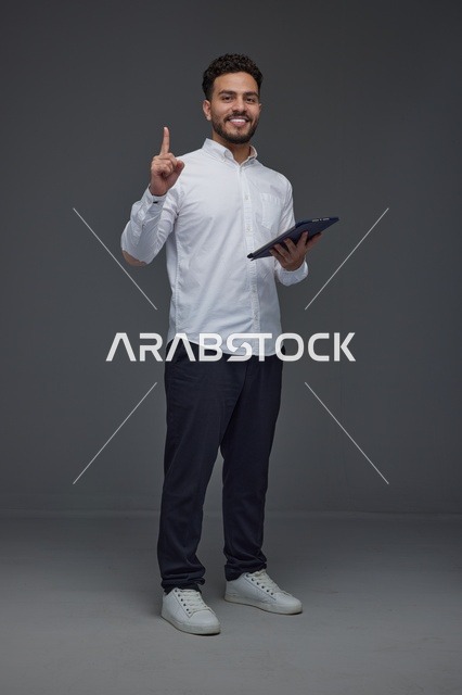 رجل عربي مبتسم يحمل جهازاً لوحياً ويشير للأعلى في الاستوديو