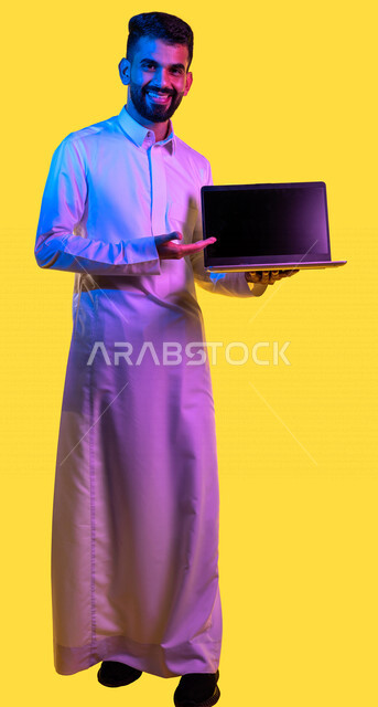 Portrait of a Saudi Arabian Gulf youth holding a laptop in his hand with facial gestures indicating happiness, businessman, professional computer and information technology jobs, e-marketing, surfing the Internet, yellow background