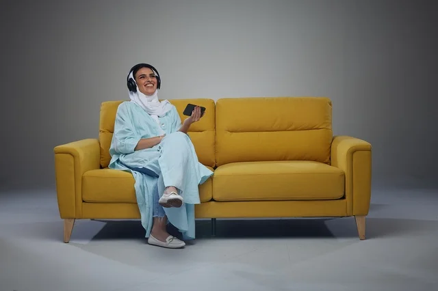 امرأة سعودية على أريكة صفراء تستخدم الهاتف والسماعات