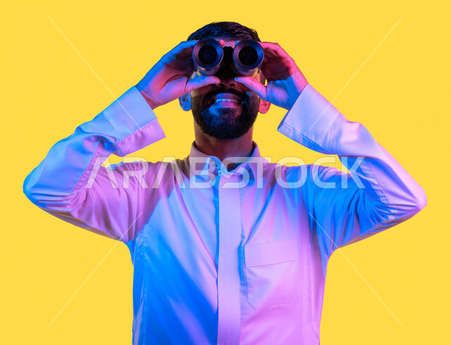 بورتريه لشاب عربي خليجي سعودي يمسك بيده ناظور و يشير بيده تجاه شئ ما رآه ، مشاهدة شيئا ما بإيماءات و تعابير وجه و يدين تدل على السعادة ، رؤية مستقبلية ، التخطيط المستقبلي ، أهداف و طموحات ، خلفية صفراء