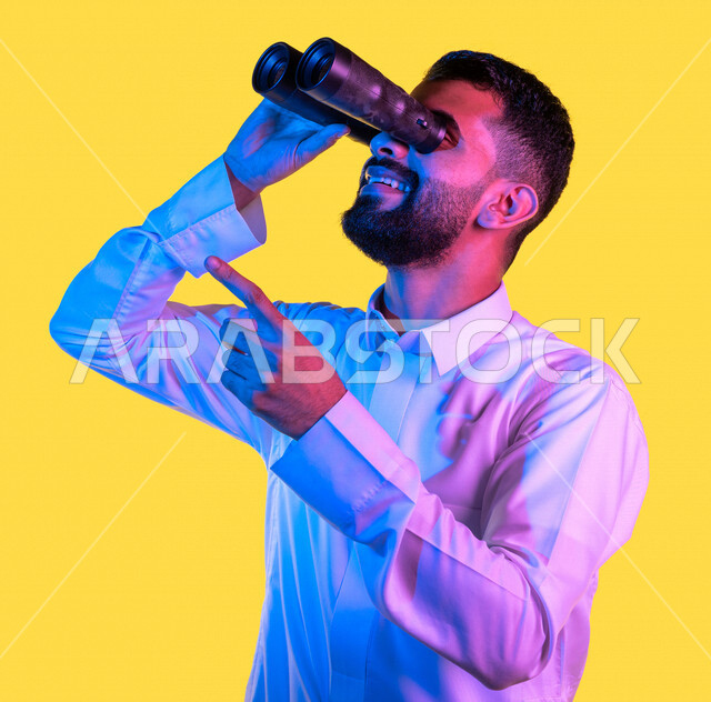 بورتريه لشاب عربي خليجي سعودي يمسك بيده ناظور و يشير بيده تجاه شئ ما رآه ، مشاهدة شيئا ما بإيماءات و تعابير وجه و يدين تدل على السعادة ، رؤية مستقبلية ، التخطيط المستقبلي ، أهداف و طموحات ، خلفية صفراء