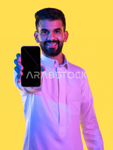 بورتريه لشاب عربي خليجي سعودي يقوم بعرض جهاز الهاتف المحمول بإيماءات تدل على السعادة ، التسويق الرقمي ، اعلانات تسويقية ترويجية ، تطبيقات الهاتف الذكي ، خلفية صفراء