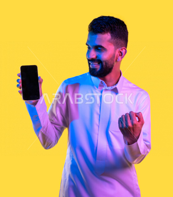 بورتريه لشاب عربي خليجي سعودي يقوم بعرض جهاز الهاتف المحمول بإيماءات تدل على السعادة و الفوز ، الحصول على الهدف المراد ، التسويق الرقمي ، اعلانات تسويقية ترويجية ، تطبيقات الهاتف الذكي ، خلفية صفراء